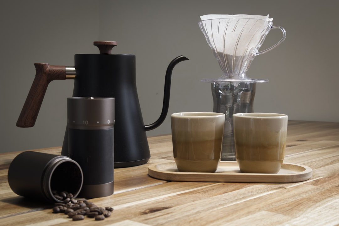 A gooseneck kettle, a V60-style pour-over dripper, a hand grinder, and two ceramic cups arranged on a wooden counter