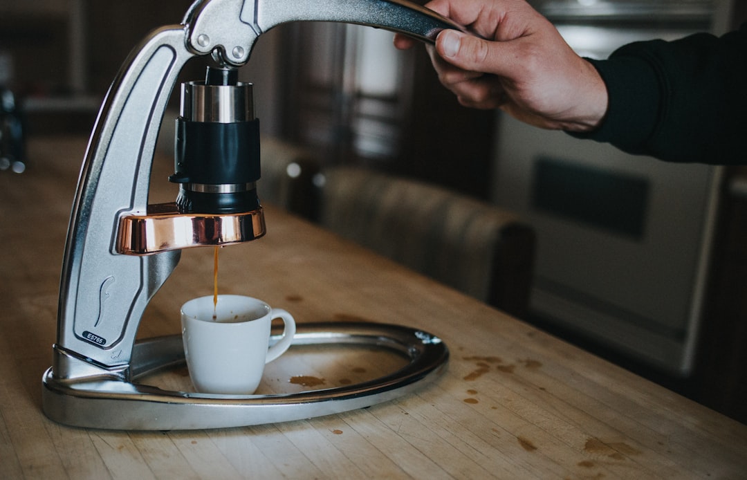 A hand pulling down the lever of a manual espresso maker as coffee streams into a small cup on a wooden counter