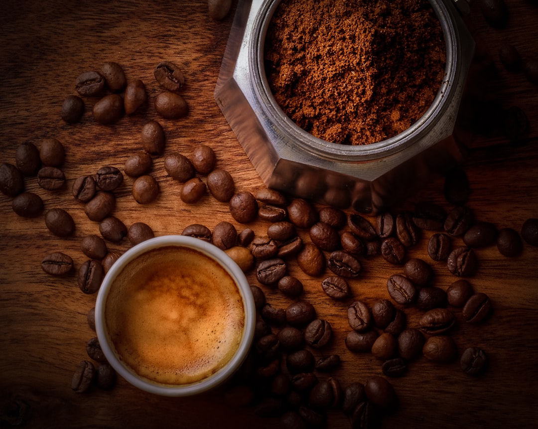A top-down perspective showing a metallic grinder hopper packed with finely milled espresso powder, adjacent to an espresso drink with crema and intact roasted beans